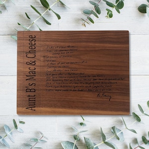 May include: A rectangular wooden cutting board with the text "Aunt B's Mac & Cheese" engraved on the left side. The board features a handwritten recipe for mac and cheese. The wood has a dark brown color.