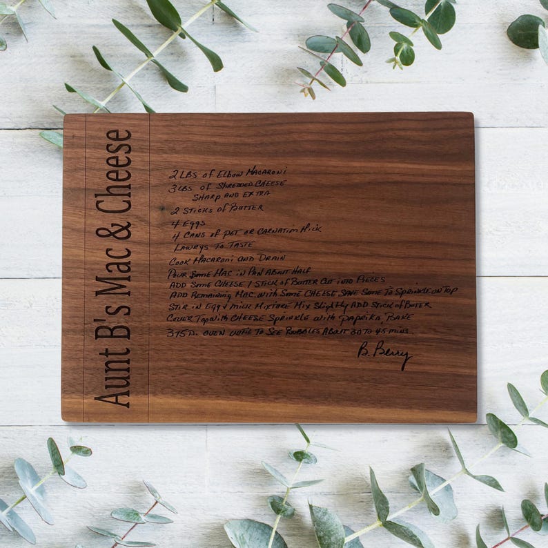 May include: A rectangular wooden cutting board with the text "Aunt B's Mac & Cheese" engraved on the left side. The board features a handwritten recipe for mac and cheese. The wood has a dark brown color.