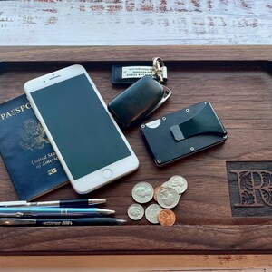 Pu&ograve; includere: Un vassoio in legno di noce scuro con vari oggetti: passaporto, smartphone, penne, monete, portafoglio e chiavi. Il vassoio ha un'iniziale intagliata e una forma rettangolare.