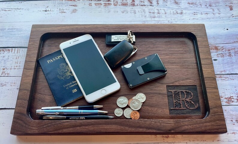 Pu&ograve; includere: Un vassoio in legno di noce scuro con vari oggetti: passaporto, smartphone, penne, monete, portafoglio e chiavi. Il vassoio ha un'iniziale intagliata e una forma rettangolare.