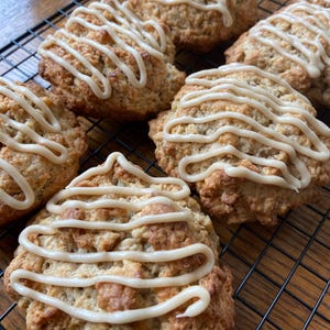 May include: Six golden-brown scones with a light-colored glaze drizzled on top, arranged on a black wire cooling rack. The baked goods have a textured surface and are freshly made.