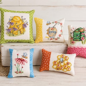May include: Five small square and rectangular fabric pillows featuring cross-stitch embroidery designs of bees, honeycombs, and hives. The pillows have colorful patterned borders in green, yellow, pink, blue, and orange, with text reading honey and be happy.