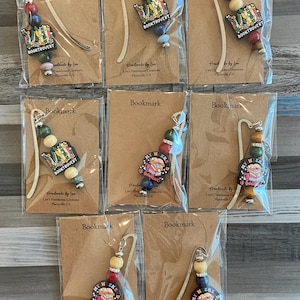 May include: A collection of handmade bookmarks. Each bookmark features a metal hook with a decorative beaded strand in various colors. The bookmarks have a charm with the text "BOOKTROVERT" or "MY WEEKEND is booked".