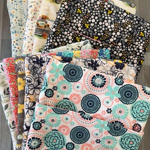 May include: A collection of folded fabric pieces featuring various patterns including floral prints, geometric circular designs, and a truck motif. The fabrics display a diverse color palette of black, white, teal, pink, yellow, and blue, arranged in a layered stack.