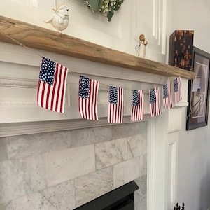 May include: A decorative bunting garland featuring seven small fabric American flags suspended on a thin brown twine string. Each flag displays the traditional red and white horizontal stripes with a blue canton containing white stars.
