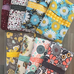 May include: A collection of eight rectangular fabric pouches featuring various patterns including floral prints with daisies, sunflowers, and abstract circles, as well as butterfly and truck motifs, each with a horizontal band across the center.
