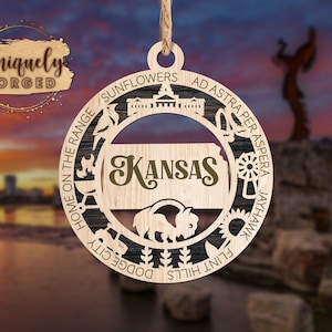 May include: A circular wooden ornament featuring a cutout map of Kansas with the word Kansas in script. The border includes icons of a building, a bird, a grill, a tractor, sunflowers, and a bison, with text reading Sunflowers, Ad Astra Per Aspera, Jayhawk, Flint Hills, Dodge City, and Home on the Range.