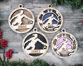 May include: Four circular wooden ornaments featuring soccer player silhouettes and soccer balls. Two ornaments are personalized with the name JONAS and two with JANICE. The outer rings are engraved with soccer terms like header, goal, and corner kick.