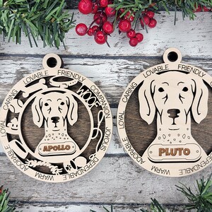 May include: Two circular wooden ornaments featuring a dog head silhouette in the center. The left ornament displays the name APOLLO and the word WOOF, while the right ornament displays the name PLUTO. Both feature engraved words like LOVABLE, FRIENDLY, COMPANION, JOYOUS, DEPENDABLE, WARM, ADORABLE, LOYAL, CARING.