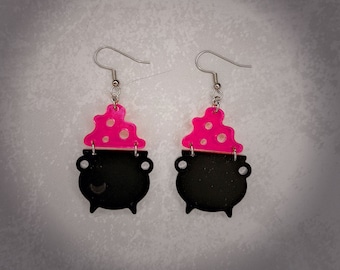 May include: A pair of black cauldron earrings with pink bubbling potion tops. The earrings are made of acrylic and have silver hooks.