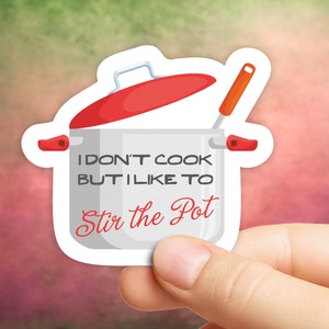Puede incluir: Una pegatina con una olla de cocina de estilo caricatura con tapa y asas rojas. El texto en la olla dice "I don't cook but I like to Stir the Pot". La pegatina se sostiene en una mano sobre un fondo borroso.
