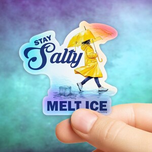 May include: A holographic sticker featuring a person in a yellow raincoat and holding a yellow and pink umbrella, walking in the rain. The sticker reads "Stay Salty Melt Ice."