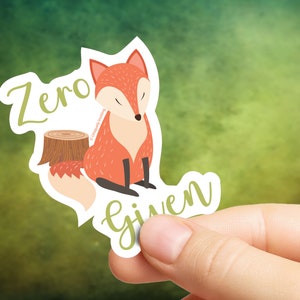 Puede incluir: Una pegatina con un zorro de dibujos animados naranja y blanco, sentado junto a un tocón de árbol. La pegatina dice "Zero Given" en cursiva verde. El zorro tiene los ojos cerrados y una postura relajada.