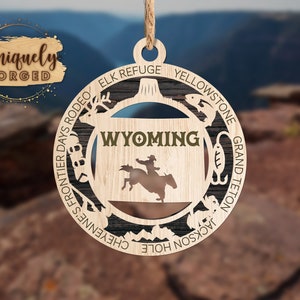 Può includere: Ciondolo in legno con lo stato del Wyoming e una silhouette di un cowboy a cavallo. Il ciondolo è decorato con varie icone che rappresentano il Wyoming, tra cui un pesce, una montagna, un cervo e un gancio. Il testo "WYOMING" è scritto in verde sul ciondolo.