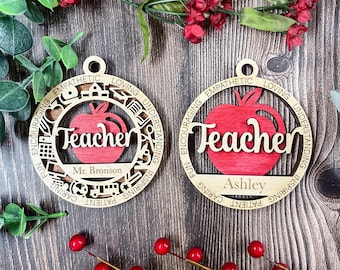Può includere: Due ornamenti circolari in legno con una mela rossa ritagliata al centro e la parola Teacher in corsivo. L'anello esterno include le parole Empathetic, Loving, Understanding, Inspiring, Patient, Caring, Fun, Encouraging e icone scolastiche.