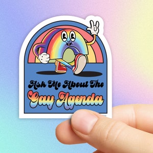 Op de afbeelding: Een sticker met een cartoon regenboogpersonage dat een vredesteken geeft. De regenboog heeft armen, benen en rode schoenen. De tekst "Ask Me About The Gay Agenda" staat eronder op een blauwe achtergrond.