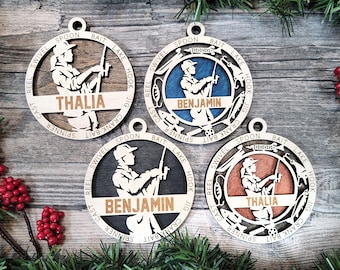 May include: Four circular wooden ornaments featuring a silhouette of a person fishing. Each ornament has a horizontal bar with the name THALIA or BENJAMIN and a border engraved with fishing terms like SWIVEL, SPOON, BAIT, LAKE, HOOK, JIG, CRANK BAIT, SPINNER, CAST, and REEL.