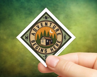 May include: A square sticker with a compass design and the words "Adventure Begins Here." The sticker features a cabin, trees, a crescent moon, and the text "Uniquely Inviting."