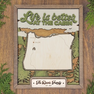 Puede incluir: Un letrero de madera con un diseño en capas que presenta un mapa de Oregón, una cabaña y árboles. El letrero dice "Life is better at the cabin" y "The Davis Family - Est. 2017".