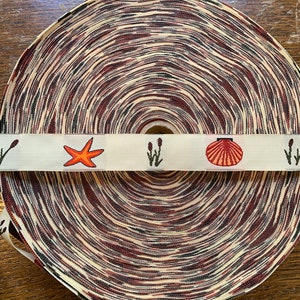 以下が含まれることがあります： 貝殻、ヒトデ、海藻のデザインが施された白いストライプのある、ベージュとブラウンの模様の入ったリボンのロールです。