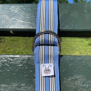 Op de afbeelding: Een blauwe en witte gestreepte stoffen riem met een zilveren gesp en een klein wit label met een cartoonbeestje.