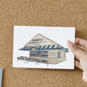 May include: A watercolour illustration of Handel's Homemade Ice Cream & Yogurt shop. The building has a tan exterior with blue and white striped awnings. The text "Handel's Homemade Ice Cream & Yogurt Since 1945" is above the awning. The image is on a corkboard.