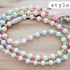Puede incluir: Un cord&oacute;n de cuentas de colores pastel con un dije de silicona rosa y un broche de metal plateado. El cord&oacute;n est&aacute; hecho de cuentas blancas con acentos de cuentas azul claro, lavanda, amarillo y rosa. El texto "style 2" es visible en la esquina superior derecha de la imagen.