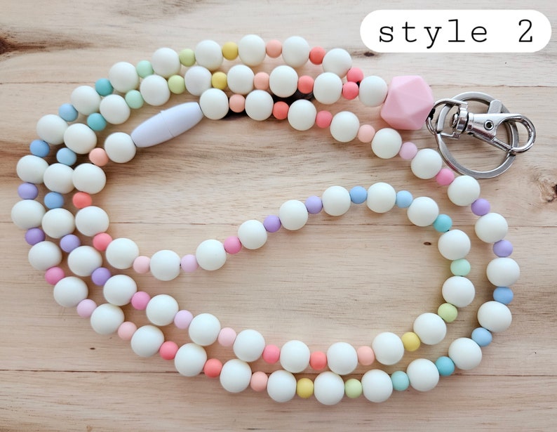 Puede incluir: Un cord&oacute;n de cuentas de colores pastel con un dije de silicona rosa y un broche de metal plateado. El cord&oacute;n est&aacute; hecho de cuentas blancas con acentos de cuentas azul claro, lavanda, amarillo y rosa. El texto "style 2" es visible en la esquina superior derecha de la imagen.