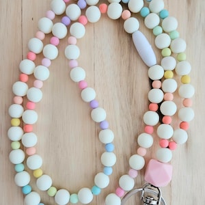 Puede incluir: Un cord&oacute;n de cuentas de colores con un dije de silicona rosa y un broche de metal plateado. Las cuentas son blancas con acentos pastel de rosa, azul, verde y morado.