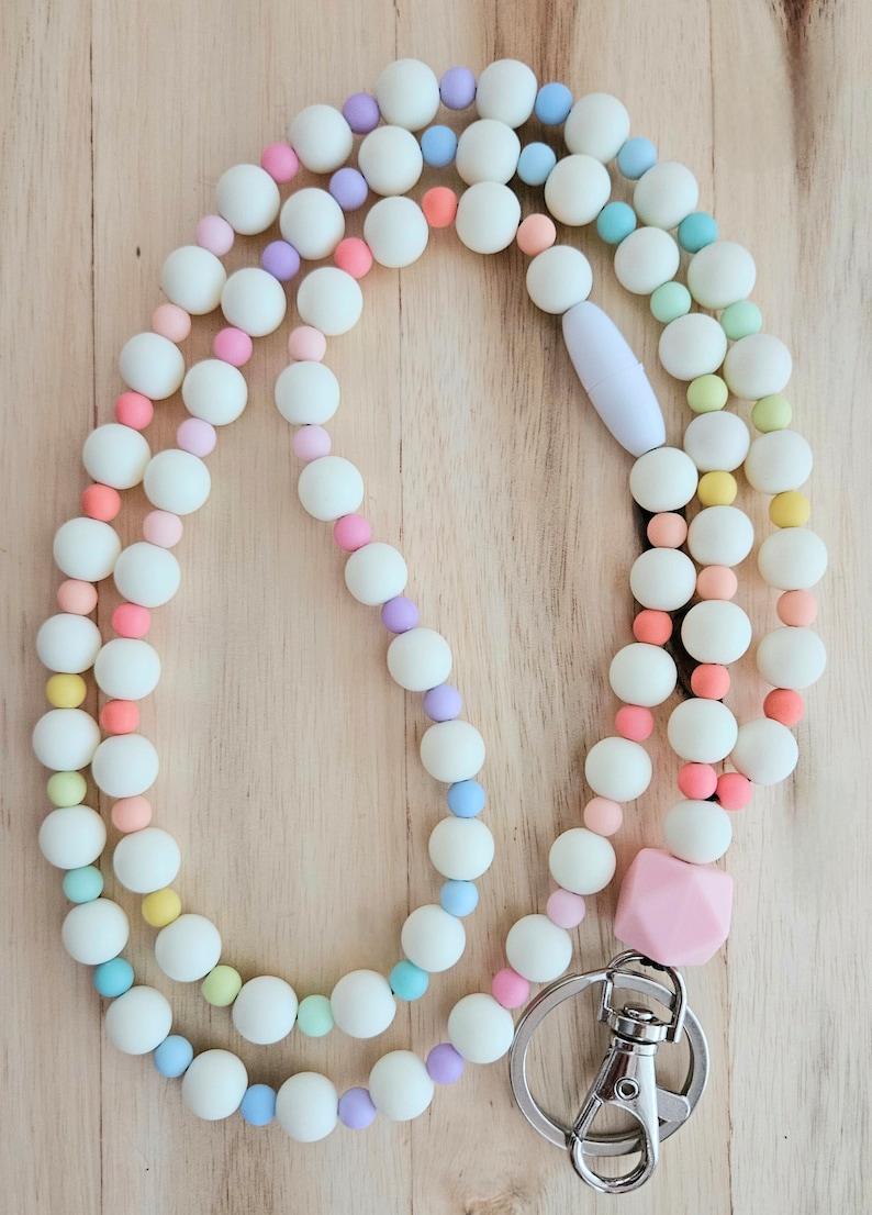 Puede incluir: Un cord&oacute;n de cuentas de colores con un dije de silicona rosa y un broche de metal plateado. Las cuentas son blancas con acentos pastel de rosa, azul, verde y morado.