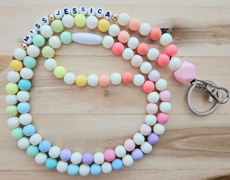 Puede incluir: Un cord&oacute;n de cuentas de silicona de color arco&iacute;ris pastel con una etiqueta de nombre blanca que dice "Miss Jessica" y un dije de silicona rosa. El cord&oacute;n tiene un broche de metal plateado.