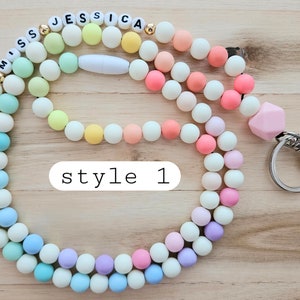 Puede incluir: Un cord&oacute;n de cuentas de colores pastel con un llavero plateado y las palabras "Miss Jessica" escritas en letras blancas. El cord&oacute;n est&aacute; hecho de dos hebras de cuentas, una en rosa pastel y la otra en amarillo y verde pastel.