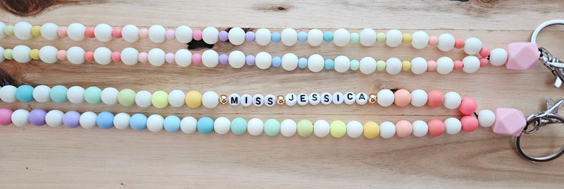 Puede incluir: Un llavero de silicona con cuentas de arco&iacute;ris pastel con un dije rosa y un llavero plateado. El llavero tiene el texto "MISS JESSICA" en letras doradas.