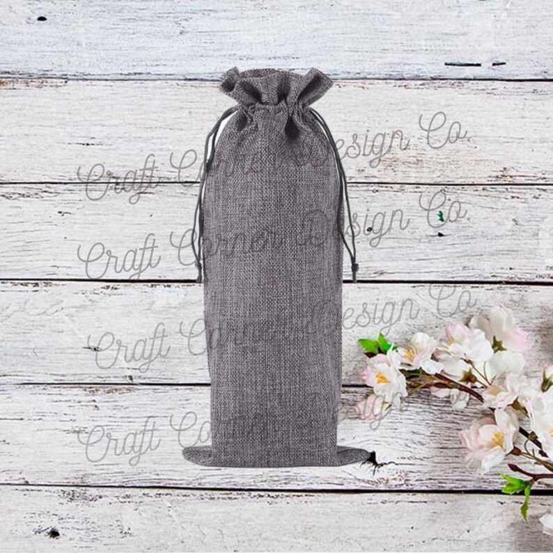 May include: A gray burlap drawstring bag for holding a bottle of wine. The bag is sitting on a white wooden surface.