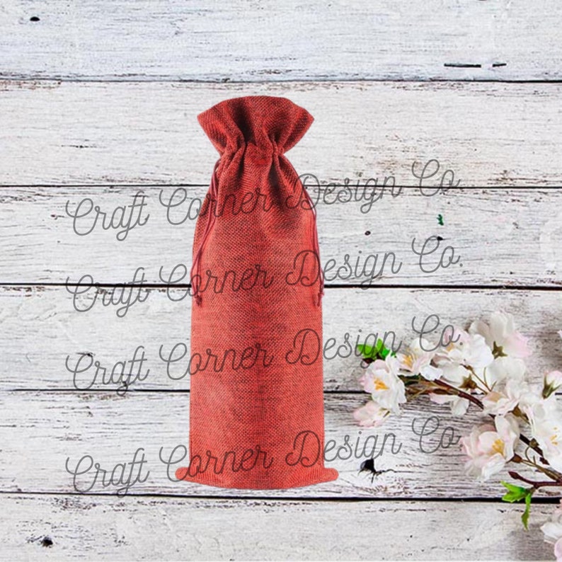 May include: A red burlap wine bottle bag with a drawstring closure. The bag is sitting on a white wooden surface.