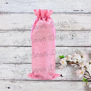 May include: Pink burlap wine bottle bag with a pink drawstring. The bag is on a white wood background.