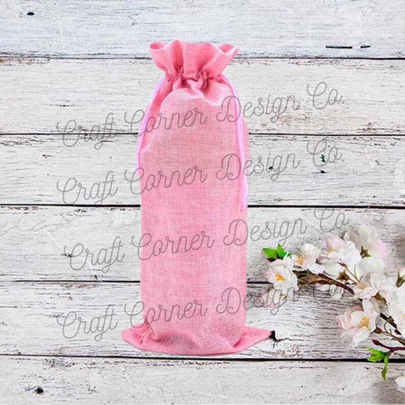 May include: Pink burlap wine bottle bag with a pink drawstring. The bag is on a white wood background.