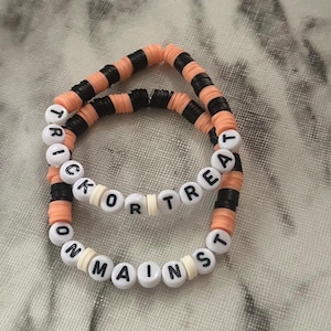 May include: Two beaded bracelets. One bracelet says "TRICK OR TREAT" in white letter beads. The other bracelet says "ON MAINST" in white letter beads. The bracelets have orange, black, and white beads.