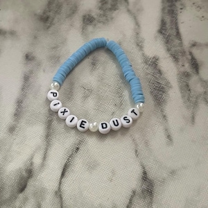 May include: A light blue beaded bracelet with the words "PIXIE DUST" spelled out in white letter beads. The bracelet features light blue disc-shaped beads and small white pearl beads. The bracelet is displayed on a light gray and white marbled surface.