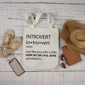 May include: White tote bag with black text that reads "INTROVERT in.tro.vert noun Just like you with a little Leave me the f*ck alone sprinkled in."