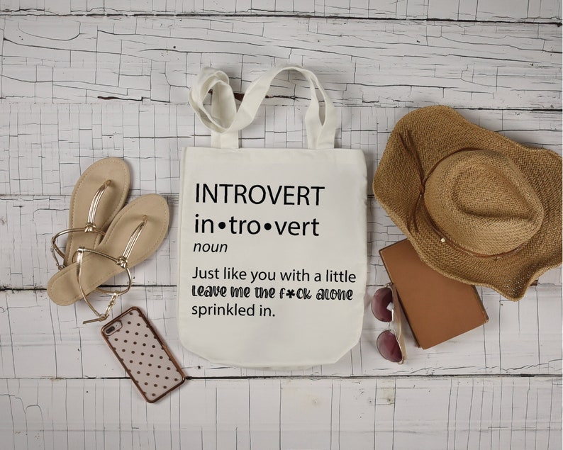 May include: White tote bag with black text that reads "INTROVERT in.tro.vert noun Just like you with a little Leave me the f*ck alone sprinkled in."