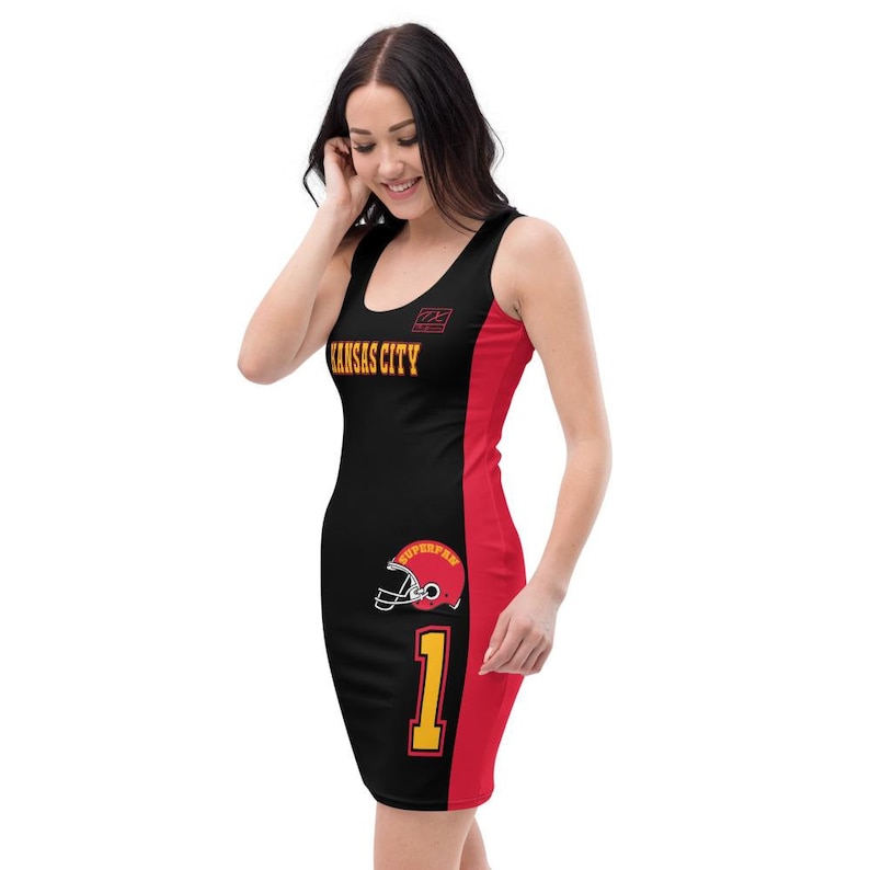 Op de afbeelding: Zwarte, mouwloze jurk met een rode streep aan de zijkant. De jurk heeft de woorden "Kansas City" en het nummer "1" in geel op de voorkant. Een rode voetbalhelm met het woord "Superfan" bevindt zich aan de zijkant van de jurk.