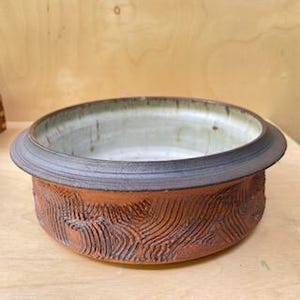 May include: A handcrafted ceramic bowl featuring a textured, reddish-brown exterior and a pale green interior. The bowl has a broad, dark rim and a circular form. The textured design on the exterior gives the piece a rustic, artisanal quality.