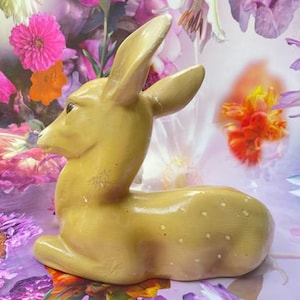 Peut inclure: Figurine de cerf en céramique jaune clair, en position assise, avec des taches blanches. Le cerf a de grandes oreilles et se détache sur un fond de fleurs colorées.