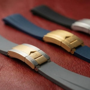 Puede incluir: Primer plano de tres correas de reloj en gris, azul y negro. Cada correa tiene un cierre metálico en oro o plata. Las correas están dispuestas sobre una superficie marrón rojiza.