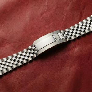 Può includere: Un cinturino per orologio in acciaio inossidabile dal design classico. Il cinturino presenta una serie di piccoli collegamenti interconnessi e una chiusura sicura. Il cinturino è esposto su una superficie in pelle rossa.
