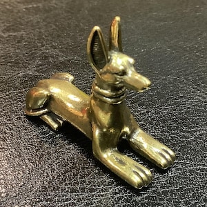 Puede incluir: Una pequeña estatua dorada de un perro en posición sentada. El perro tiene orejas grandes y erguidas y lleva un collar. La estatua descansa sobre una superficie oscura y texturizada.