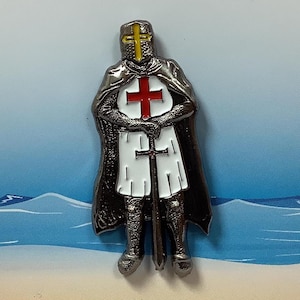 May include: A silver pin badge featuring a knight in armour with a red cross on his chest. The knight is holding a sword and wearing a helmet with a yellow cross. The badge says "Pin Badge" and "Made in the U.K."