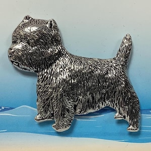 May include: A silver-colored metal pin of a West Highland White Terrier dog standing on a blue and white background.