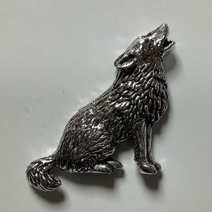 May include: A silver-colored metal wolf figurine in a howling pose. The wolf is detailed with textured fur and is sitting upright with its head tilted back and mouth open. The figurine is on a white surface.